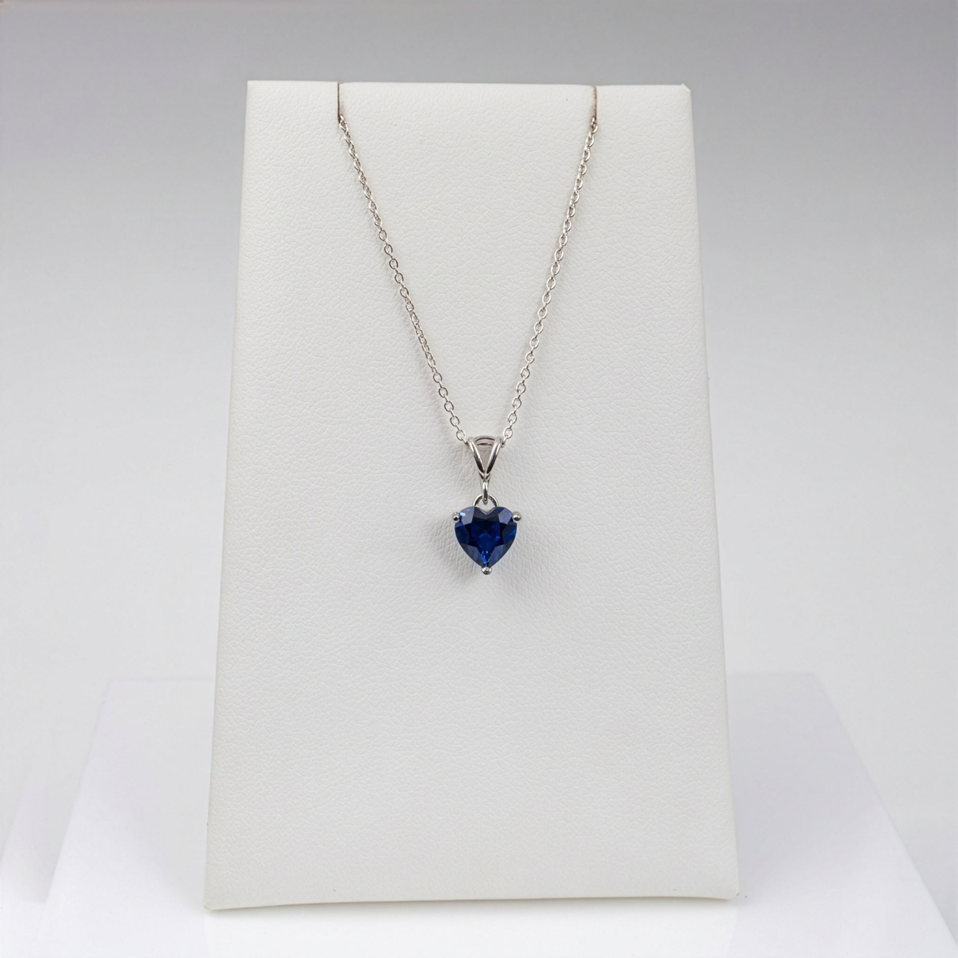 A silver pendant necklace with a heart-shaped blue lab sapphire gemstone, displayed on a white jewelry stand against a light gray background.