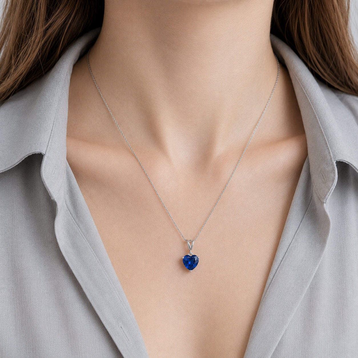 A close-up of a model wearing a delicate silver chain necklace with a heart-shaped blue gemstone pendant, styled with a light gray collared shirt.