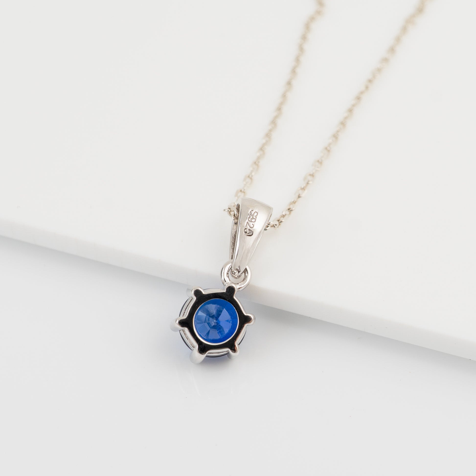 A silver and lab sapphire  pendant necklace featuring a round blue gemstone in a prong setting, showing the reverse side and resting on a clean white surface.