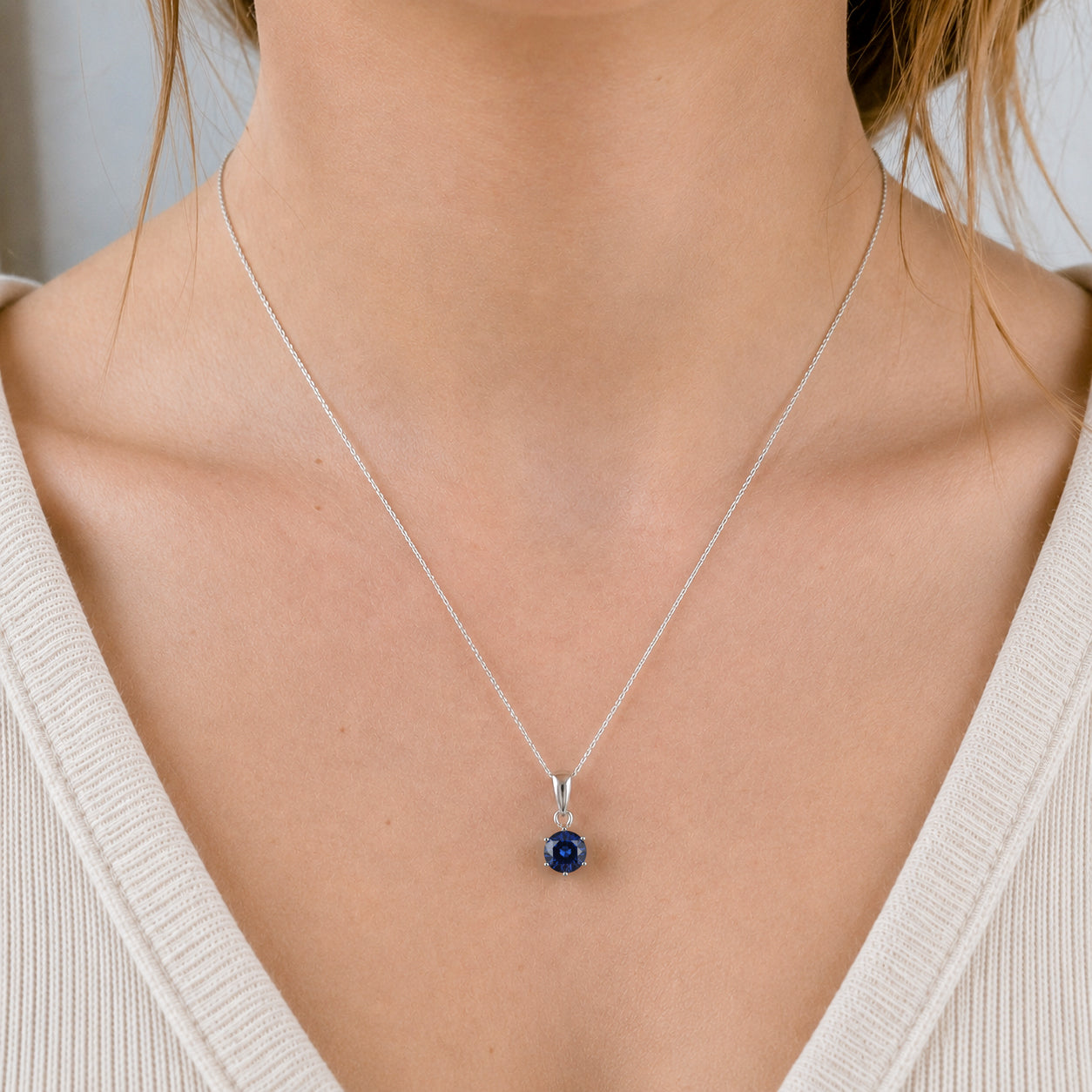 A close-up of a model's neck and chest wearing a delicate silver chain necklace with a small round lab blue sapphire gemstone pendant.