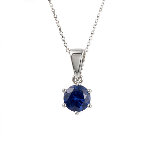A silver and lab sapphire pendant necklace featuring a round-cut deep blue gemstone in a four-prong setting, suspended from a delicate silver chain against a plain white background.