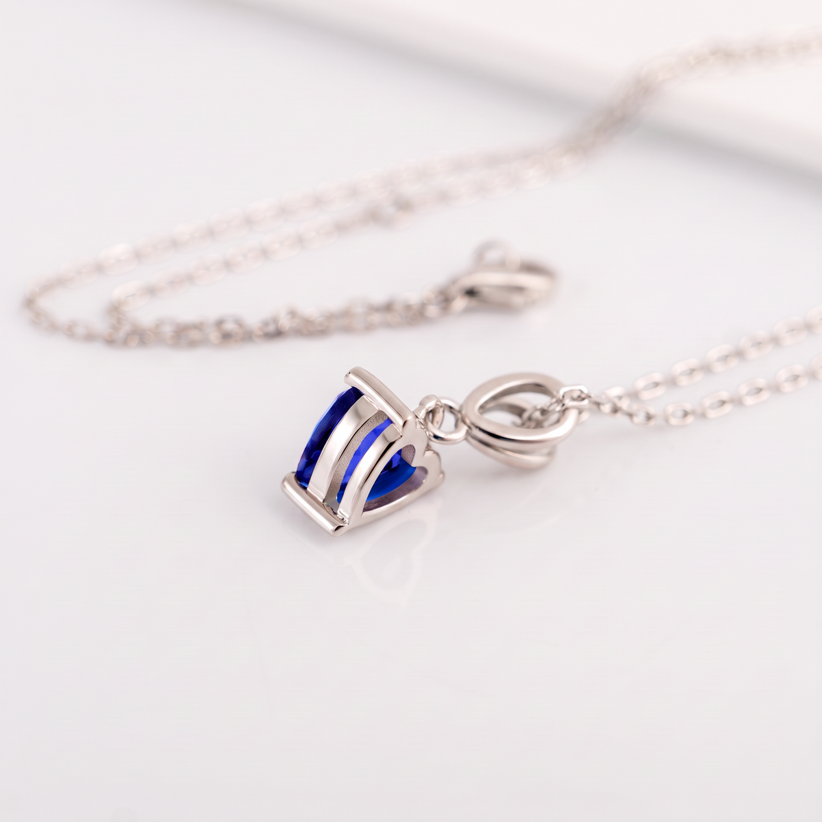 A silver necklace with a small royal blue sapphire heart pendant photographed on a clean white surface with the chain softly blurred in the background.