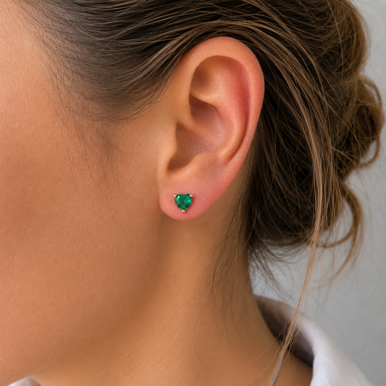 A close-up of a woman's ear wearing a small heart-shaped green gemstone stud earring in a silver setting.