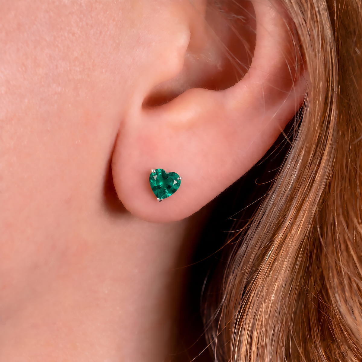 A close-up of a person's ear wearing a small heart-shaped lab emerald gemstone stud earring.