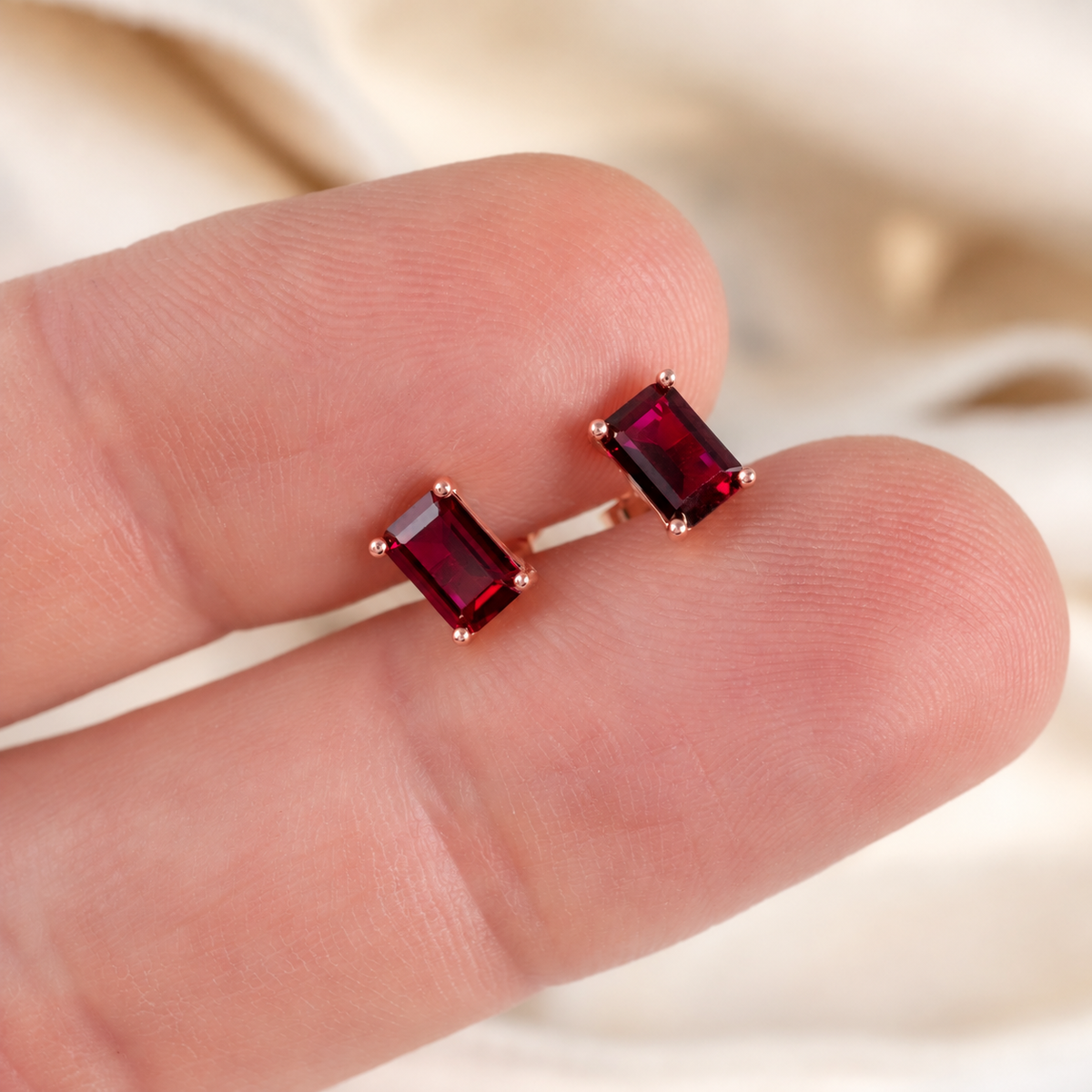 A pair of rectangular lab grown gemstone stud earrings in a rose gold setting, shown resting on a person's fingers against a soft beige fabric background.