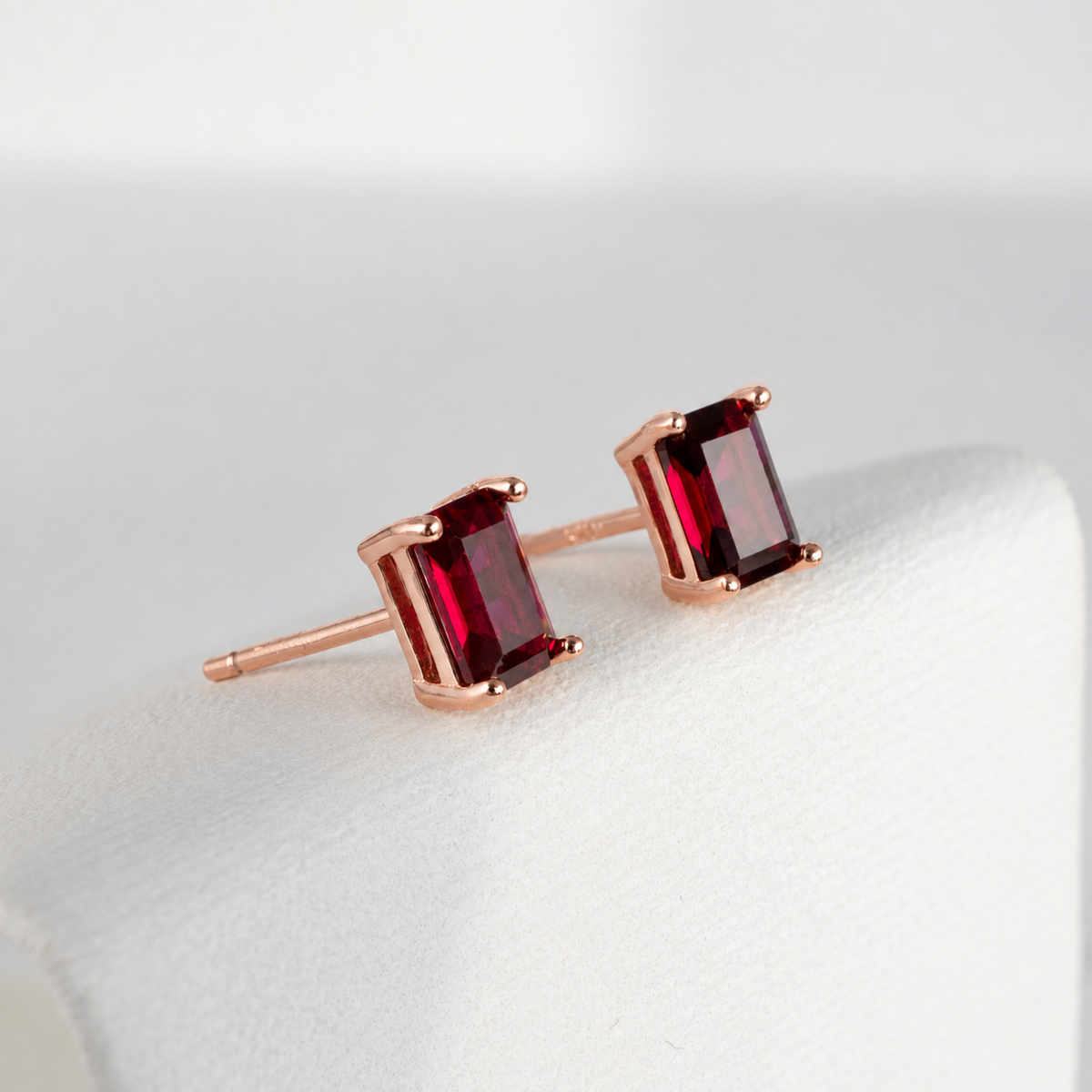 A pair of rose gold stud earrings with rectangular emerald-cut lab grown ruby gemstones, displayed on a white jewelry stand 