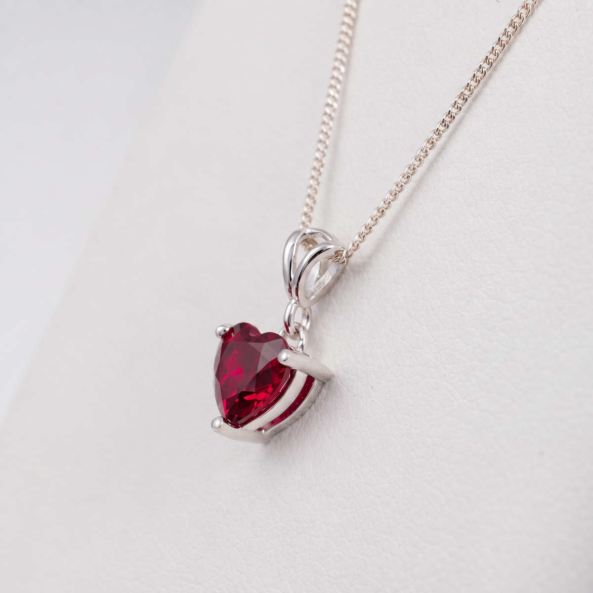 A silver heart-shaped pendant necklace with a faceted lab ruby gemstone, displayed on a clean white background.