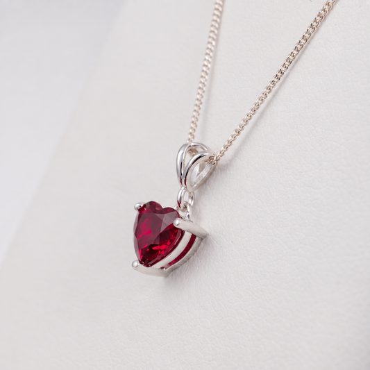 A silver heart-shaped pendant necklace with a faceted lab ruby gemstone, displayed on a clean white background.