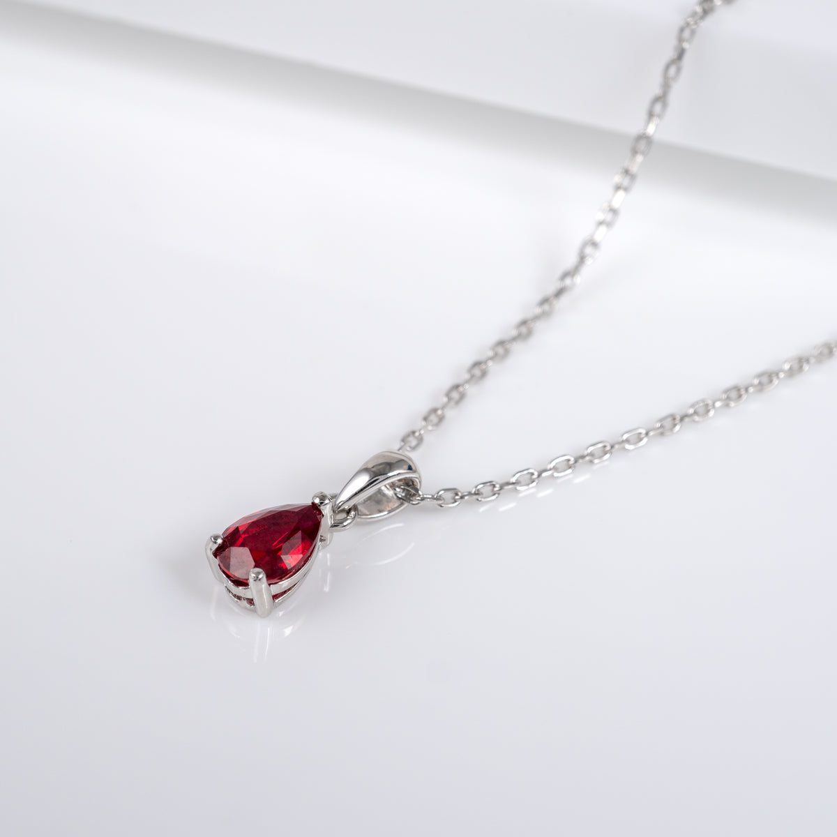 A silver-toned chain necklace with a teardrop-shaped lab ruby gemstone pendant, displayed on a clean white surface.