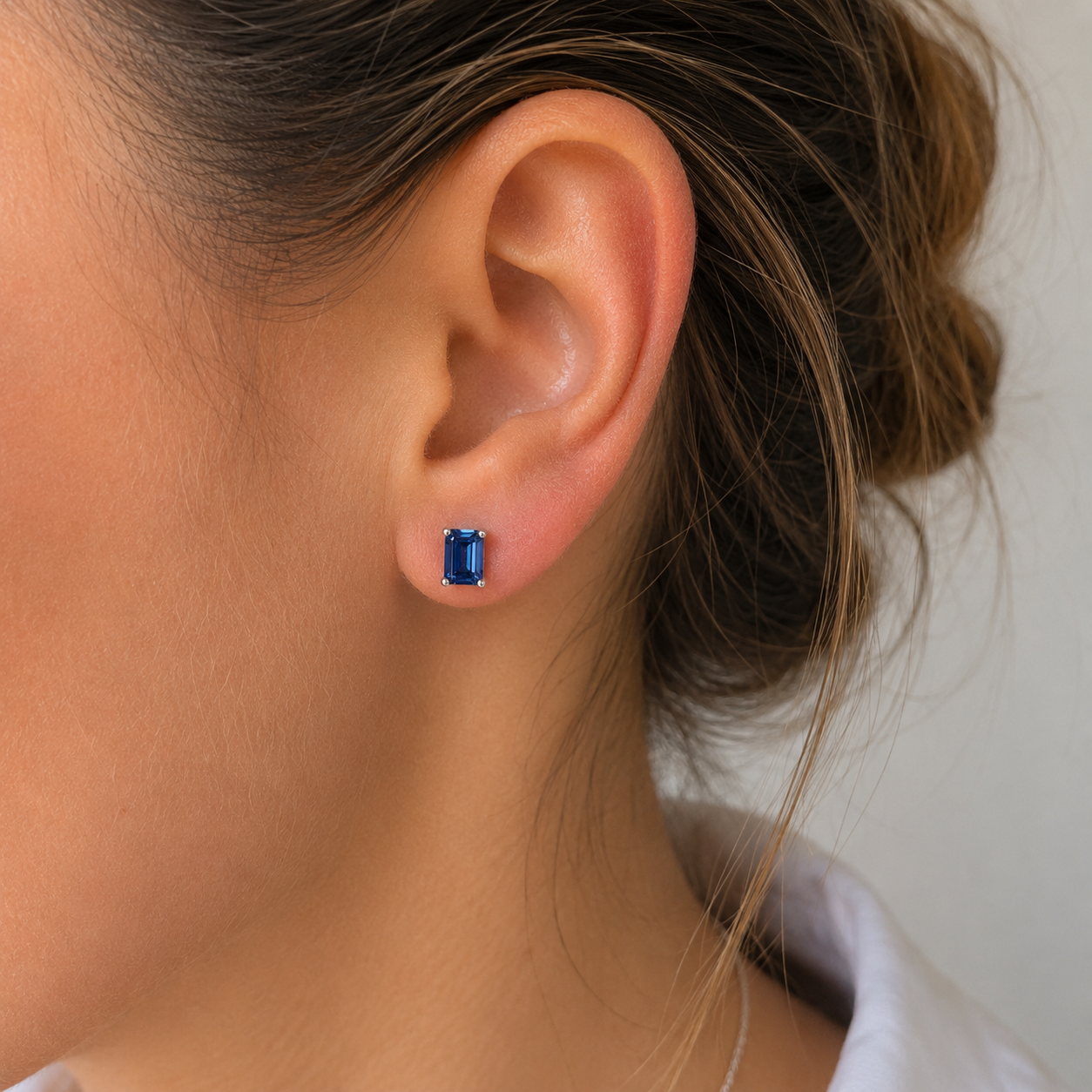 A close-up of a woman's ear wearing a small rectangular blue gemstone stud earring in a silver setting.