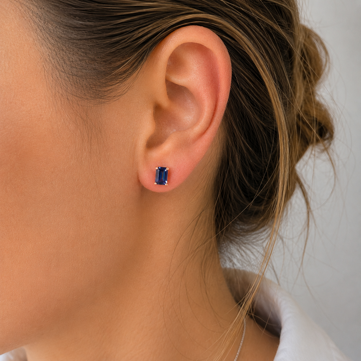 A close-up of a woman's ear wearing a small rectangular blue gemstone stud earring in a silver setting.