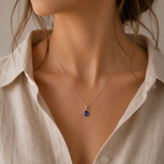 A close-up of a woman wearing a delicate silver chain necklace with a teardrop-shaped blue lab sapphire gemstone pendant, styled with a light beige linen shirt.