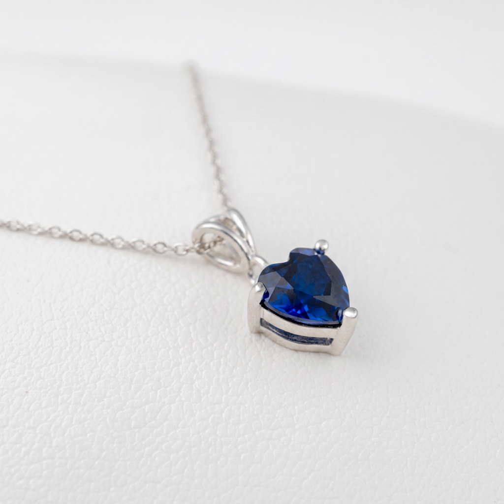 A silver heart-shaped pendant necklace with a rich blue lab sapphire blue faceted gemstone, resting on a textured white surface.