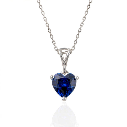 A heart-shaped lab sapphire gemstone pendant necklace in a silver-toned prong setting on a delicate silver chain, shown against a white background with a soft reflection below.