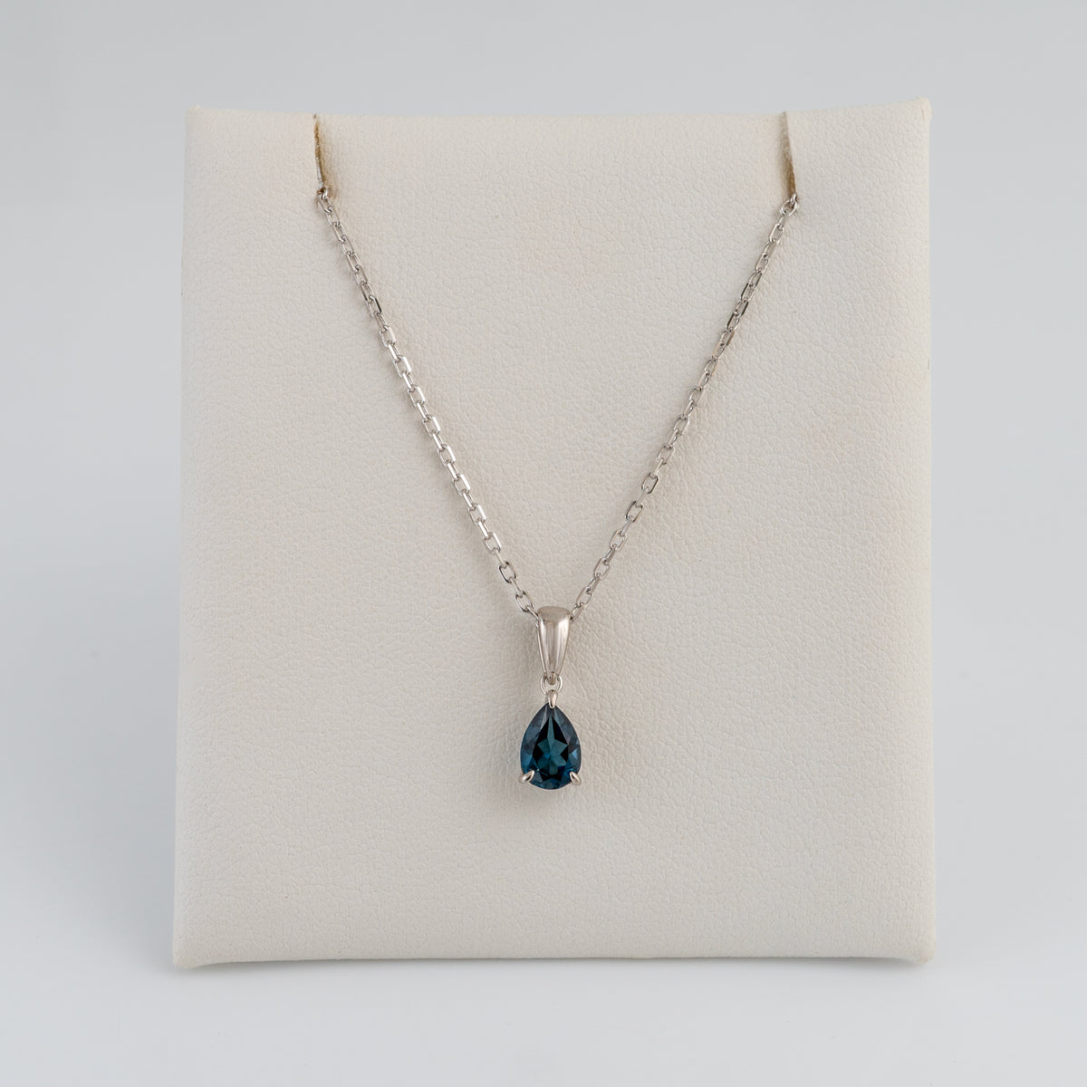 A silver chain necklace with a teardrop-shaped london blue topaz gemstone pendant, displayed on a cream-colored jewelry stand against a light gray background.