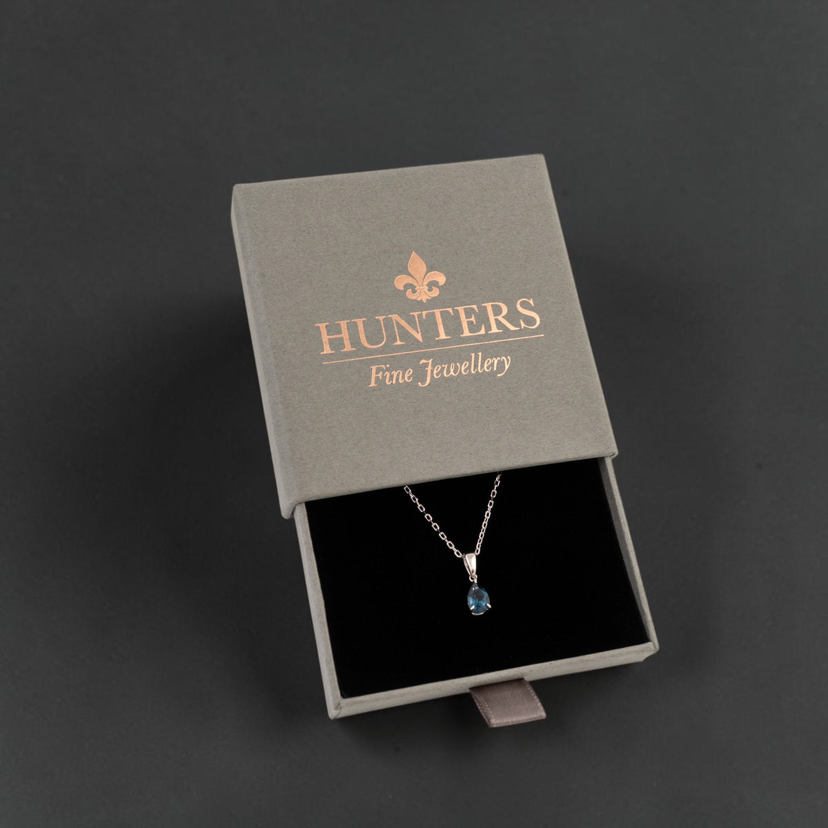 A silver pendant necklace with a small london blue topaz teardrop gemstone displayed inside an open gray Hunters Fine Jewellery gift box on a dark grey background.
