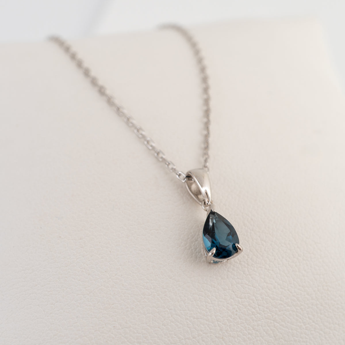 A silver chain necklace with a teardrop-shaped london blue topaz gemstone pendant, displayed on a textured off-white surface.