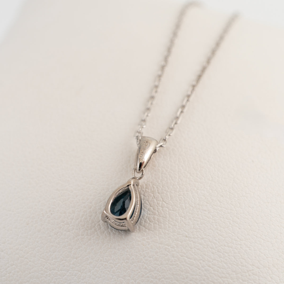 Reverse view of a London blue topaz teardrop necklace in sterling silver, showing the open-backed gemstone setting and tapered bail on a white cushion.