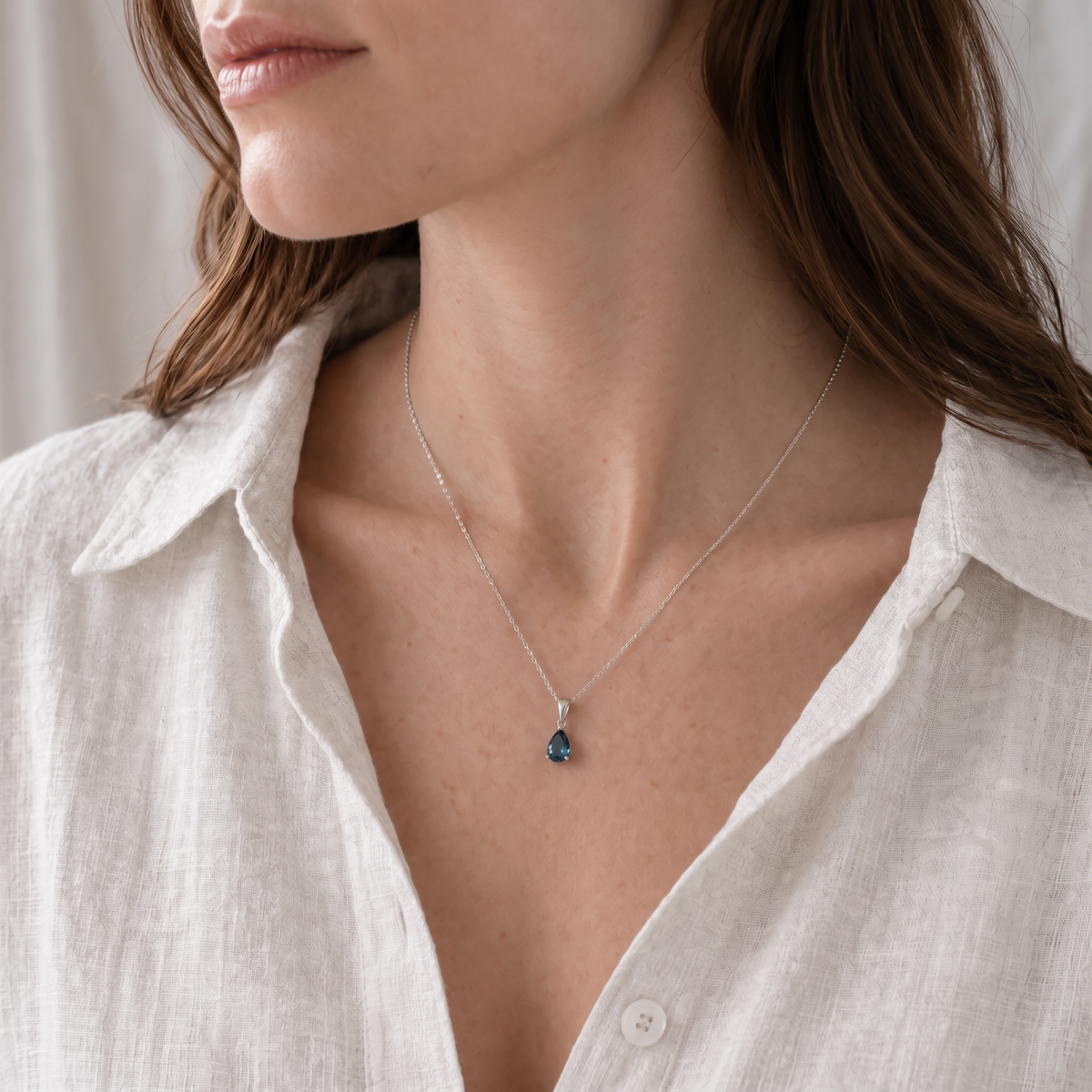 A close-up of a woman wearing a delicate silver chain necklace with a small teardrop-shaped london blue topaz gemstone pendant, styled with a white linen shirt.