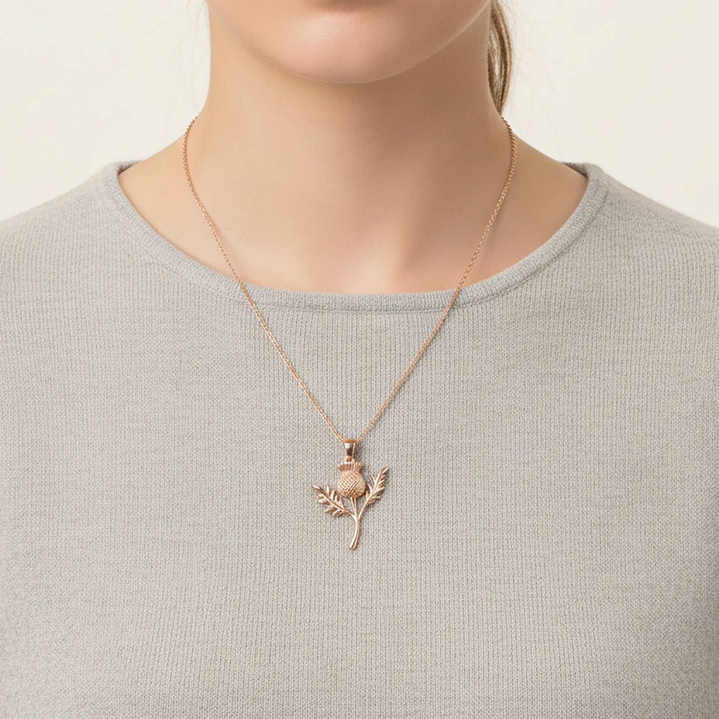 Person wearing a gold necklace with a thistle pendant on a plain background
