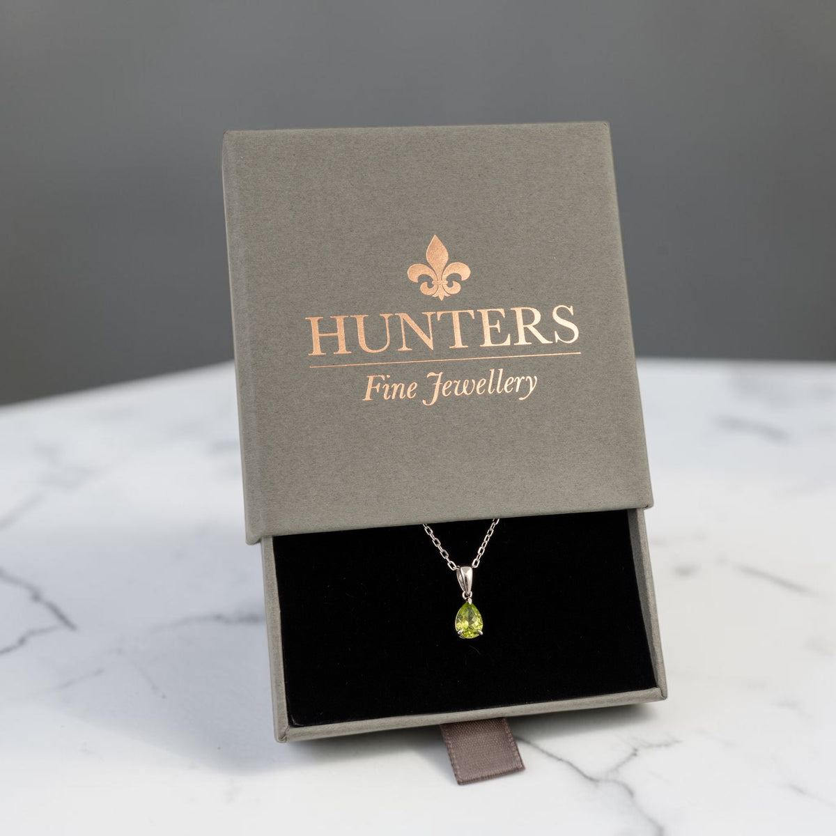 Natural peridot teardrop necklace in sterling silver displayed in a Hunters Fine Jewellery presentation box on a white marble-patterned surface.