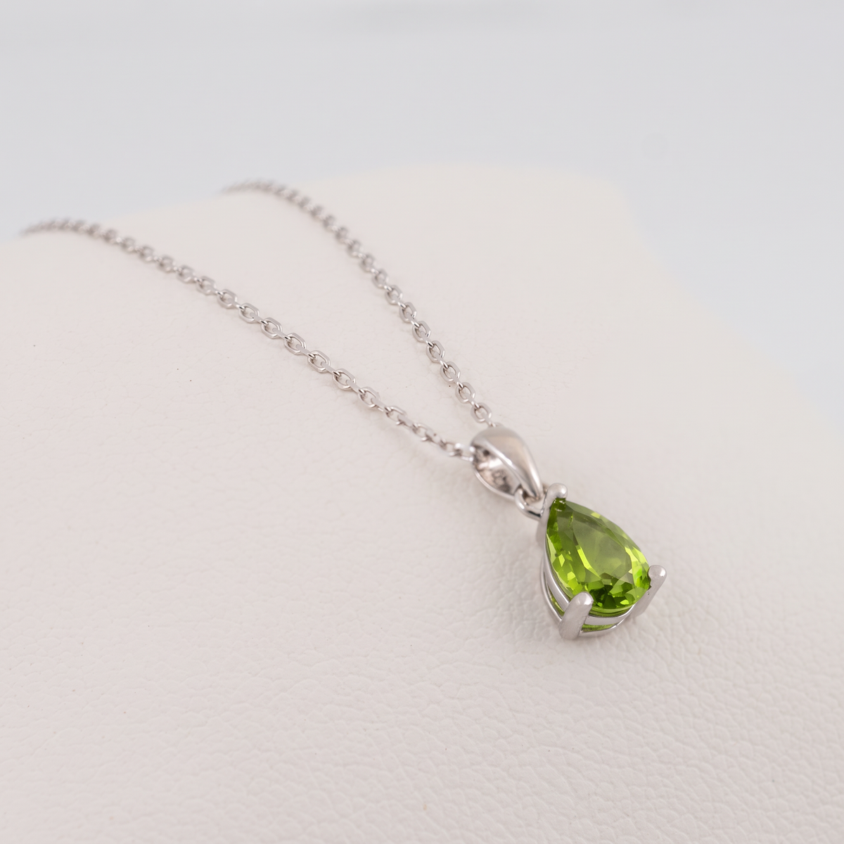 Natural peridot teardrop necklace in sterling silver resting at a right-facing angle on a white cushion, showing the green gemstone facets and claw setting.