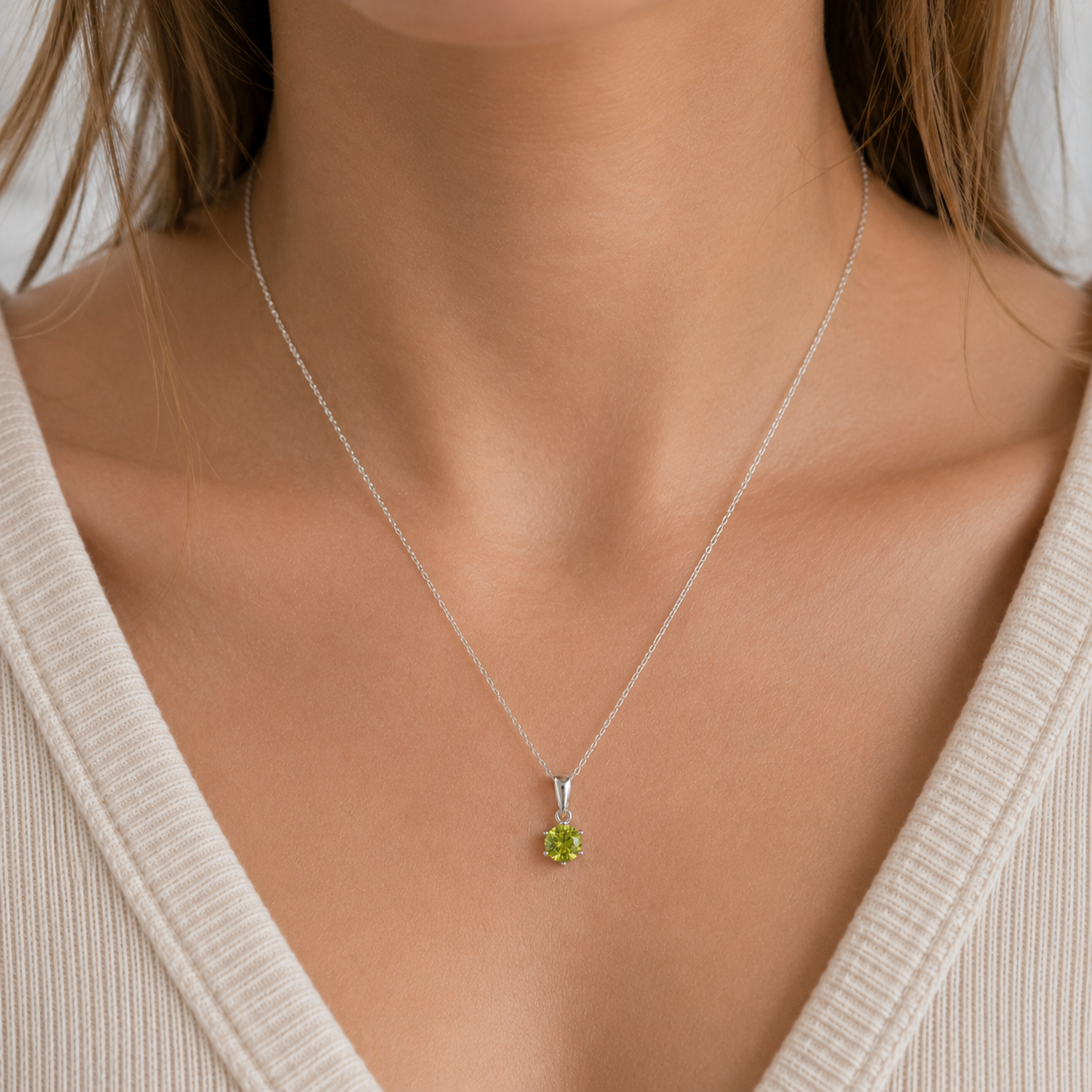 A close-up of a person's neck and chest wearing a delicate silver chain necklace with a small green gemstone pendant, paired with a cream-colored ribbed V-neck top.