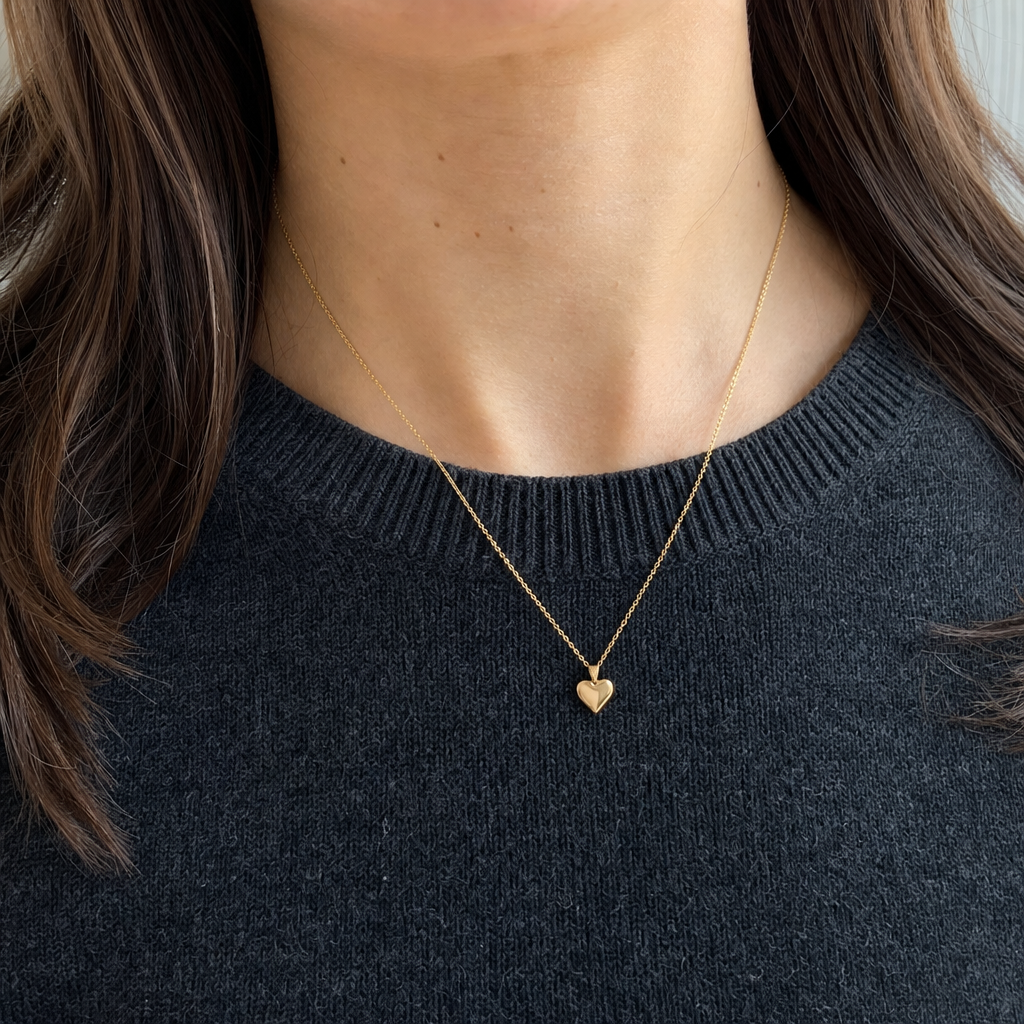 A close-up of a person wearing a delicate gold chain necklace with a small heart pendant, styled with a dark gray knit sweater.