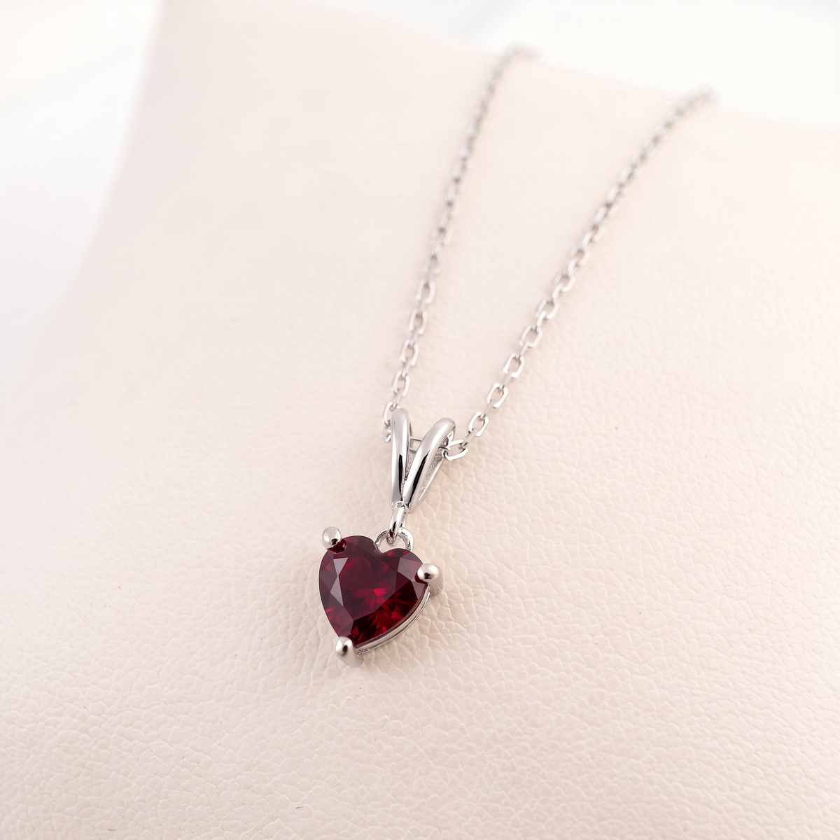 A heart-shaped pigeon blood ruby pendant in a silver setting on a delicate silver chain, displayed on a light beige textured surface.