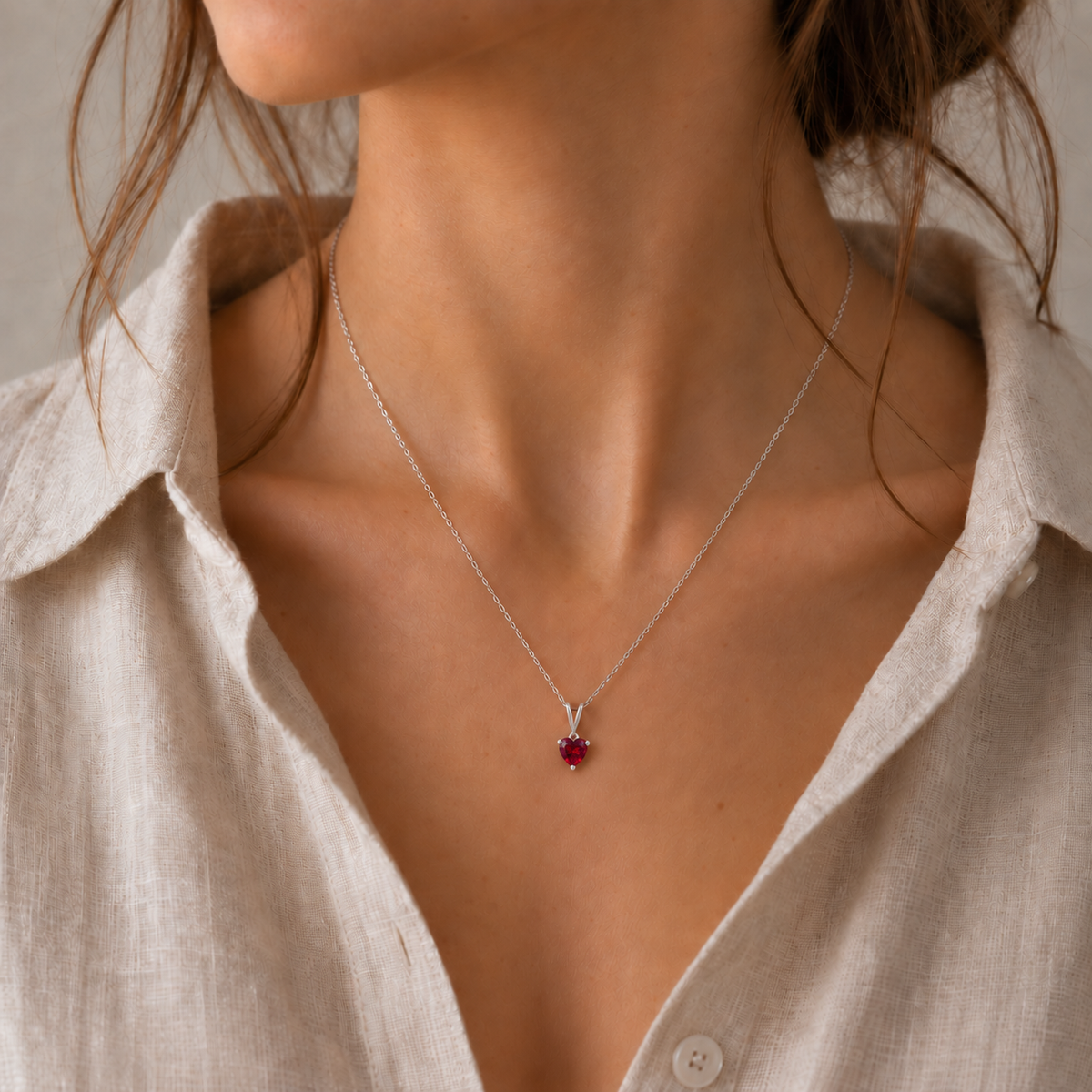 A close-up of a model wearing a delicate silver chain necklace with a small heart-shaped red gemstone pendant, styled with a light beige linen shirt.