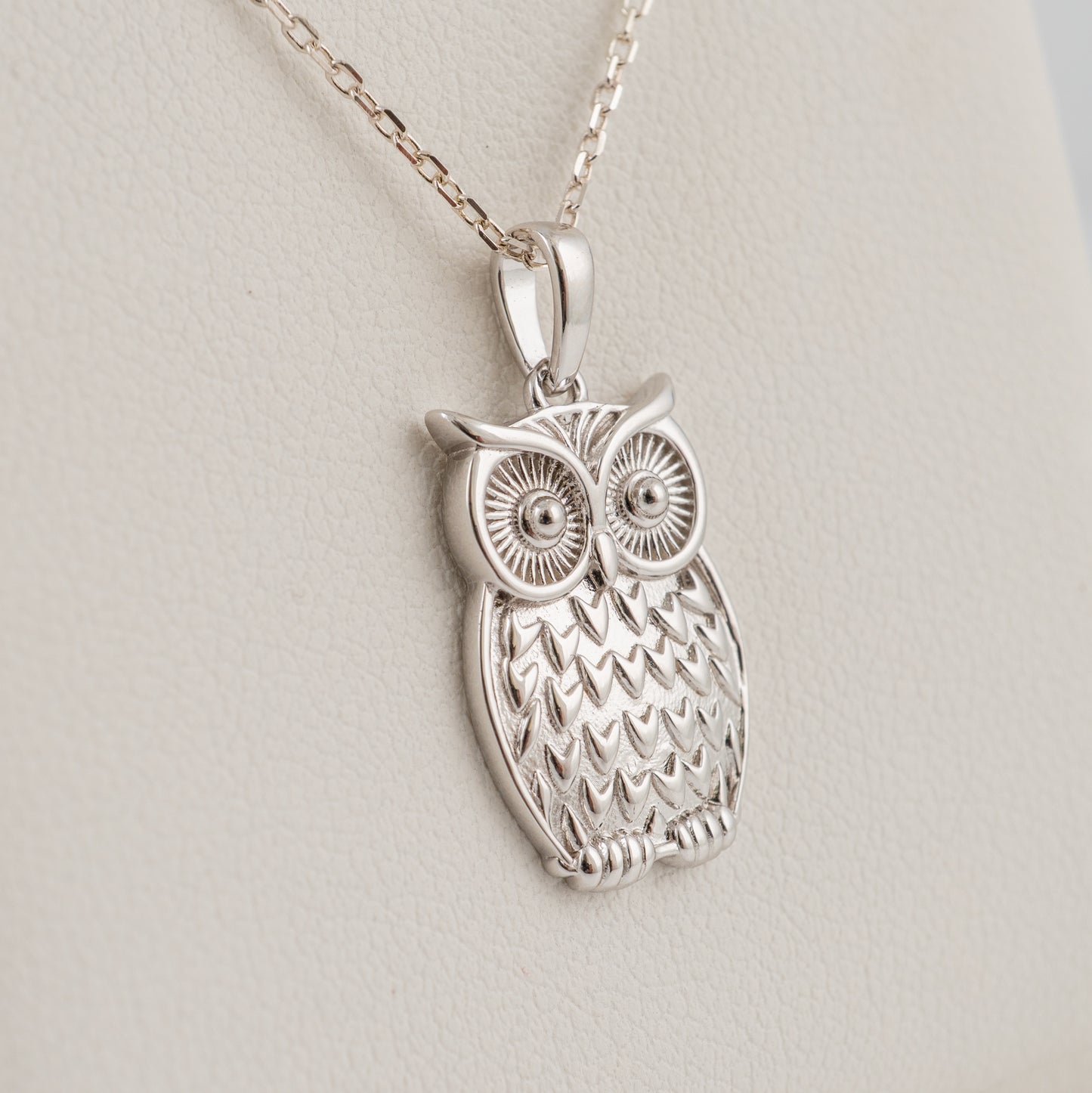 A plain silver owl pendant necklace with a textured feather pattern and large detailed eyes, hanging from a silver chain against a light beige textured background.