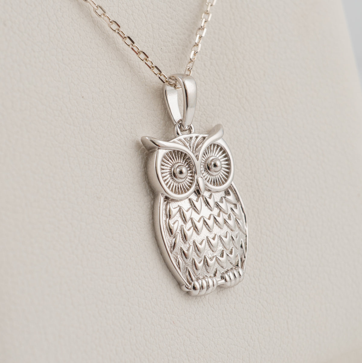 A plain silver owl pendant necklace with a textured feather pattern and large detailed eyes, hanging from a silver chain against a light beige textured background.