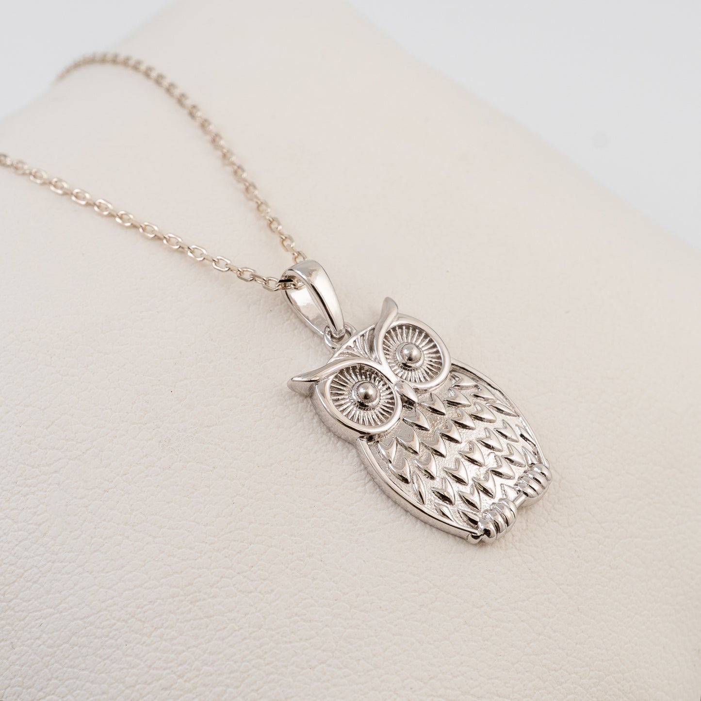 A plain silver owl pendant necklace with a detailed textured body and large round eyes, hanging from a delicate silver chain on a cream-colored display stand.