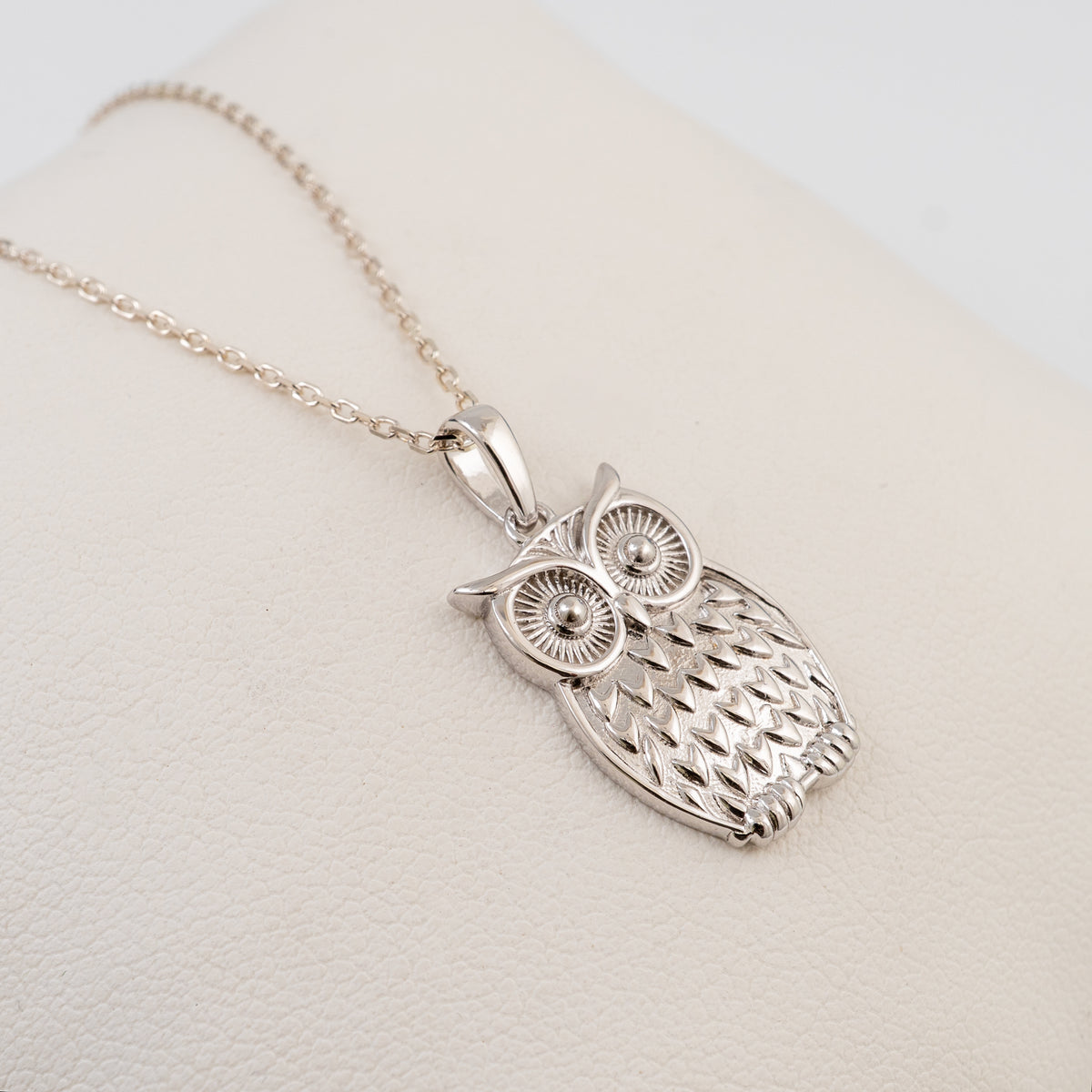 A plain silver owl pendant necklace with a detailed textured body and large round eyes, hanging from a delicate silver chain on a cream-colored display stand.