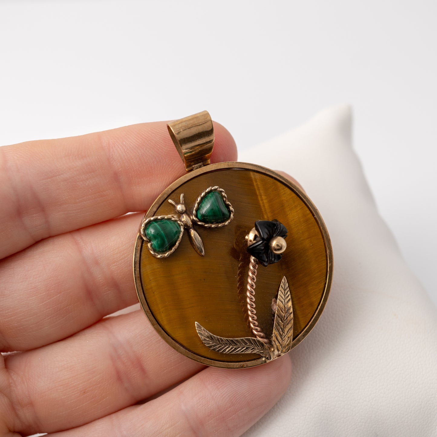 Hand holding a vintage 9ct gold butterfly pendant with tiger's eye background and malachite details, showcasing its size and scale.