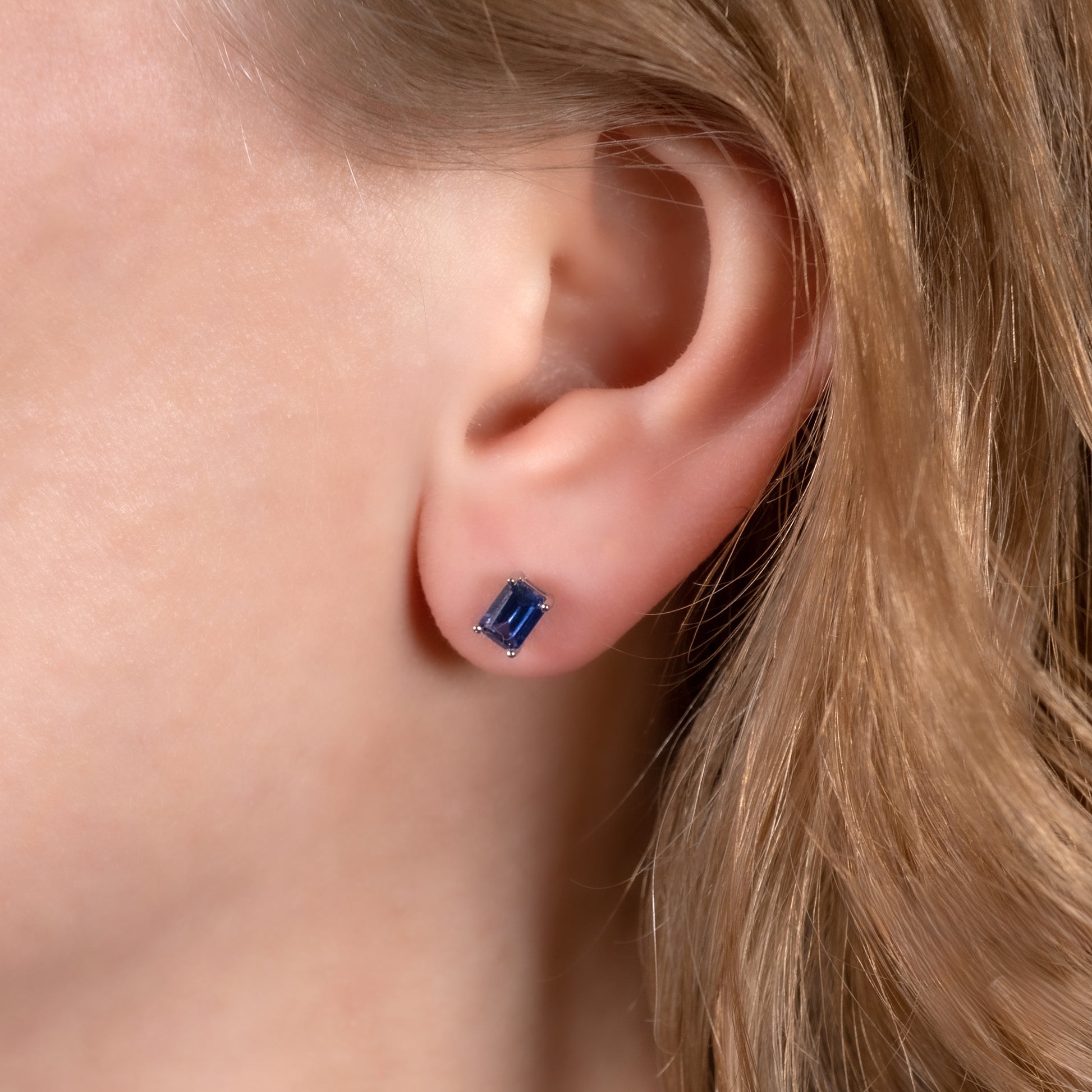 A close-up of a person's ear wearing a small rectangular blue sapphire gemstone stud earring.