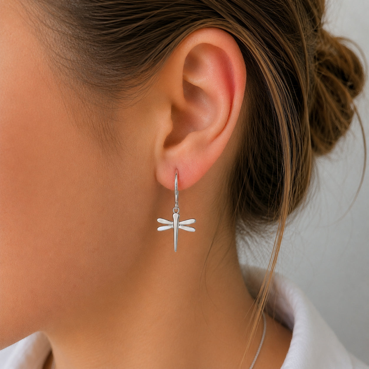 A close-up of a woman's ear wearing a delicate silver dragonfly dangle earring, with her hair tied back in a bun and a white top visible.