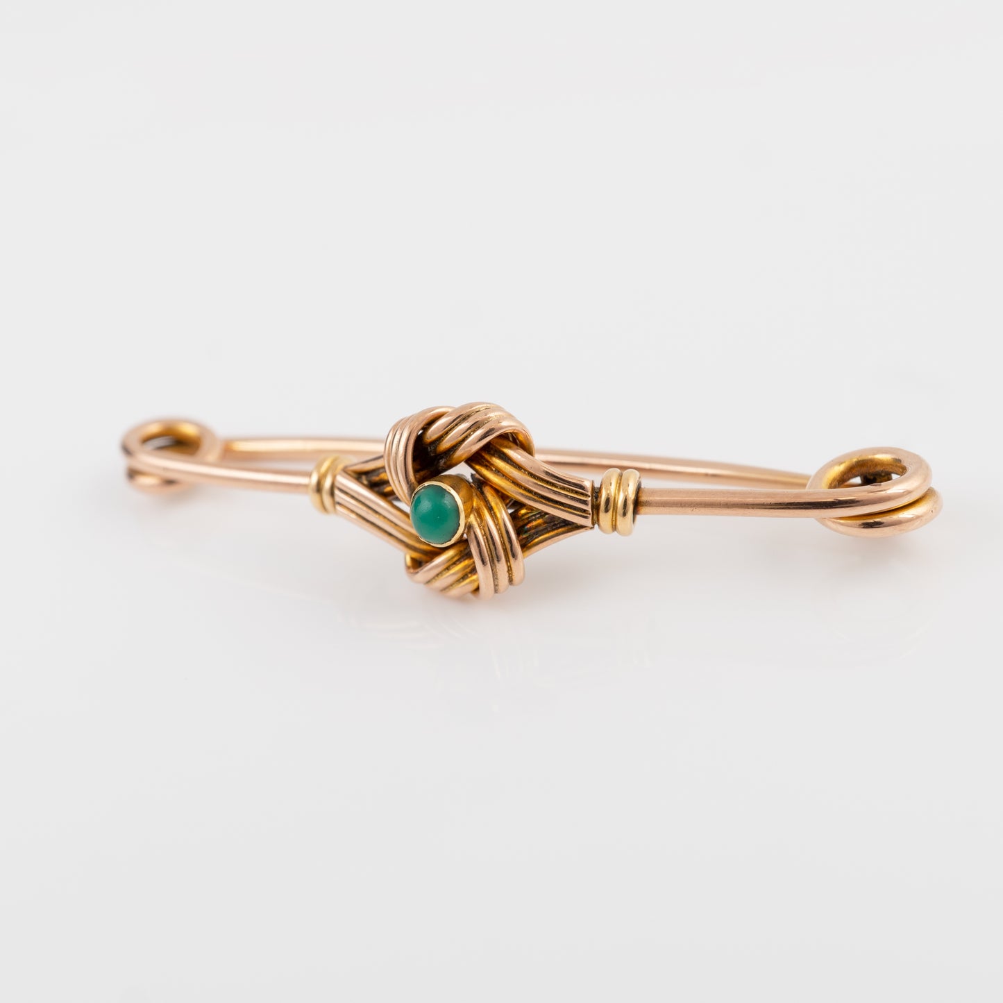 A gold brooch featuring a central woven knot with a small round green stone accent, set against a clean white background.