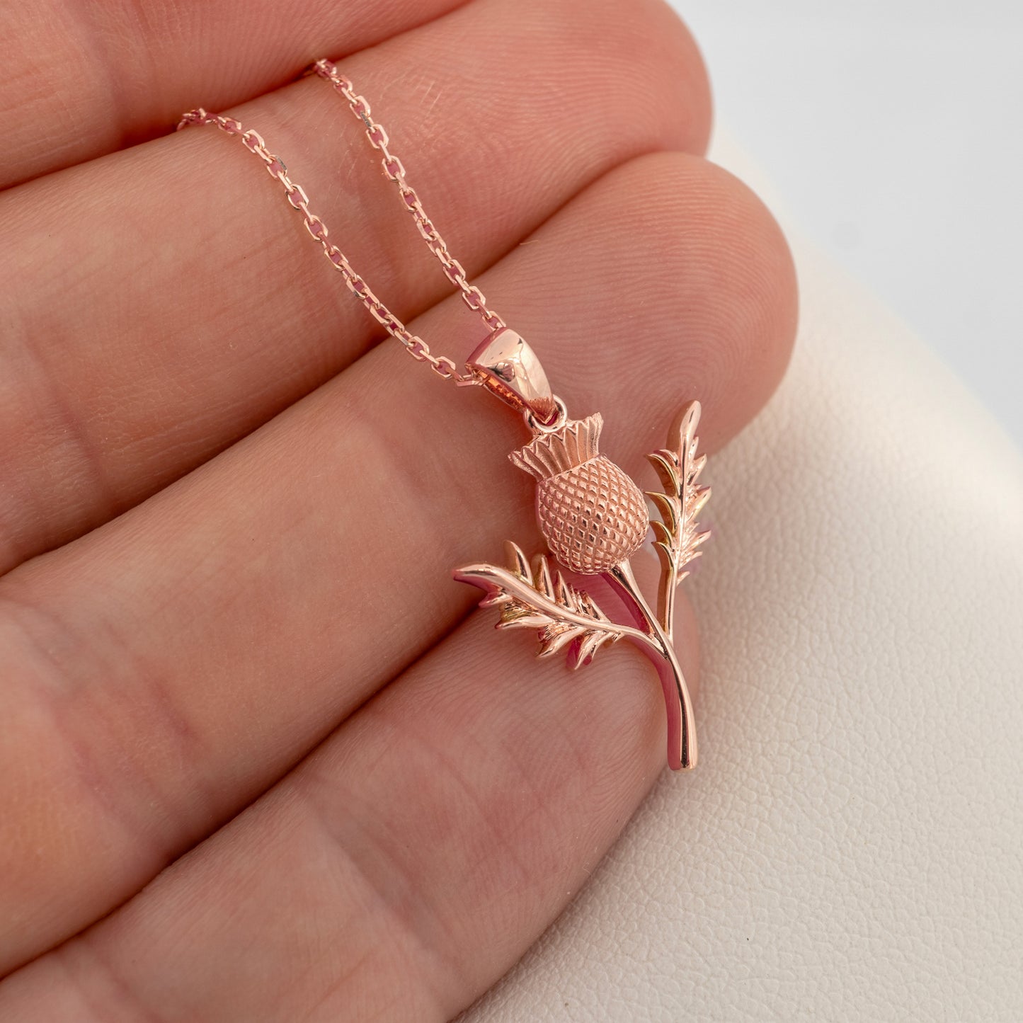 Rose gold thistle pendant necklace on a hand against a neutral background