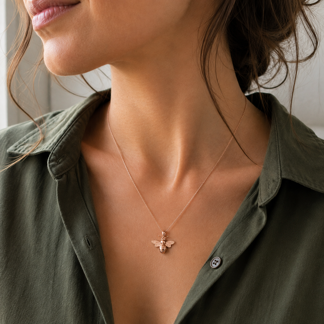 A close-up lifestyle shot of a woman wearing a delicate rose gold bee pendant necklace on a fine chain, paired with an olive green button-down shirt.