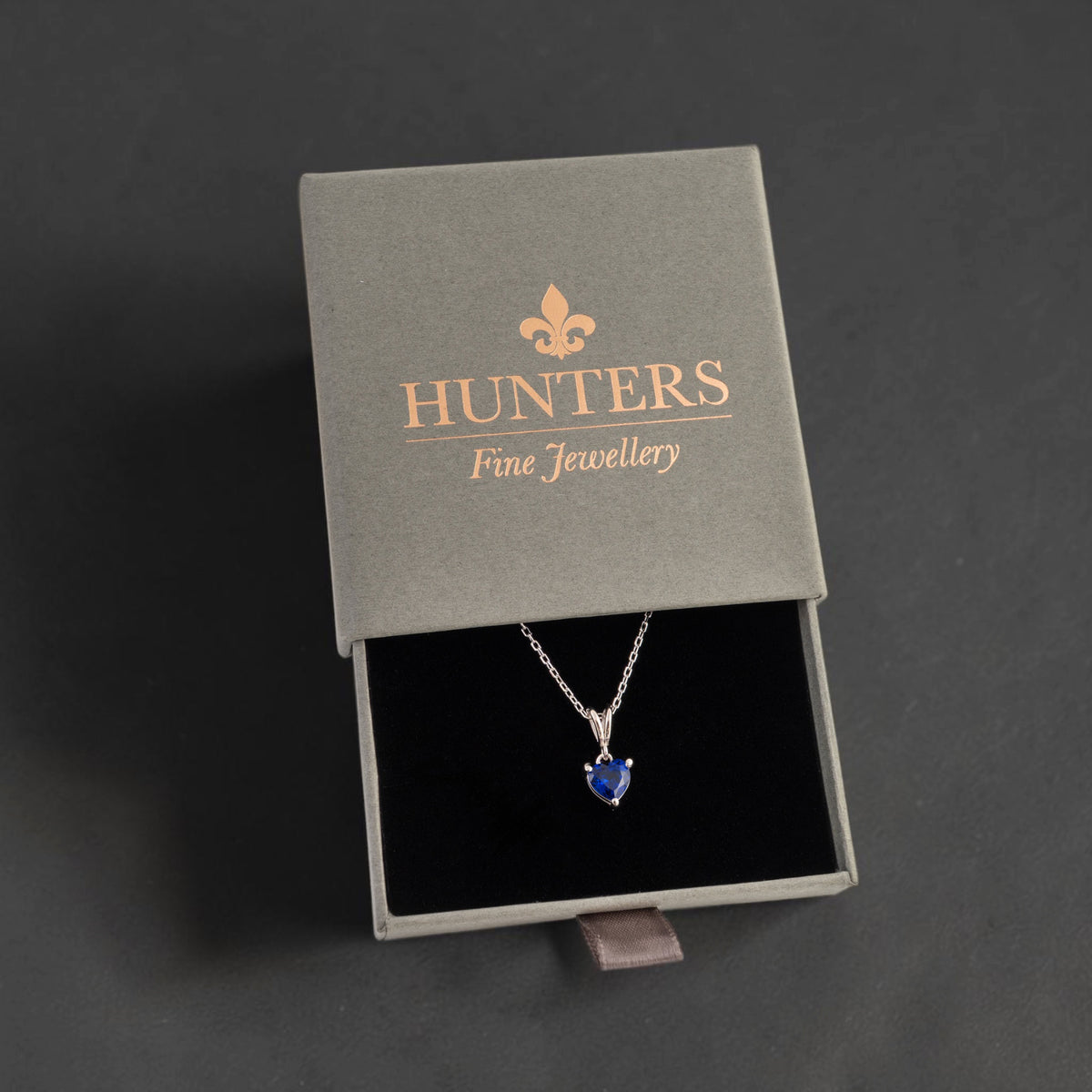 A silver pendant necklace with a heart-shaped green gemstone displayed inside an open gray Hunters Fine Jewellery gift box on a dark grey background.
