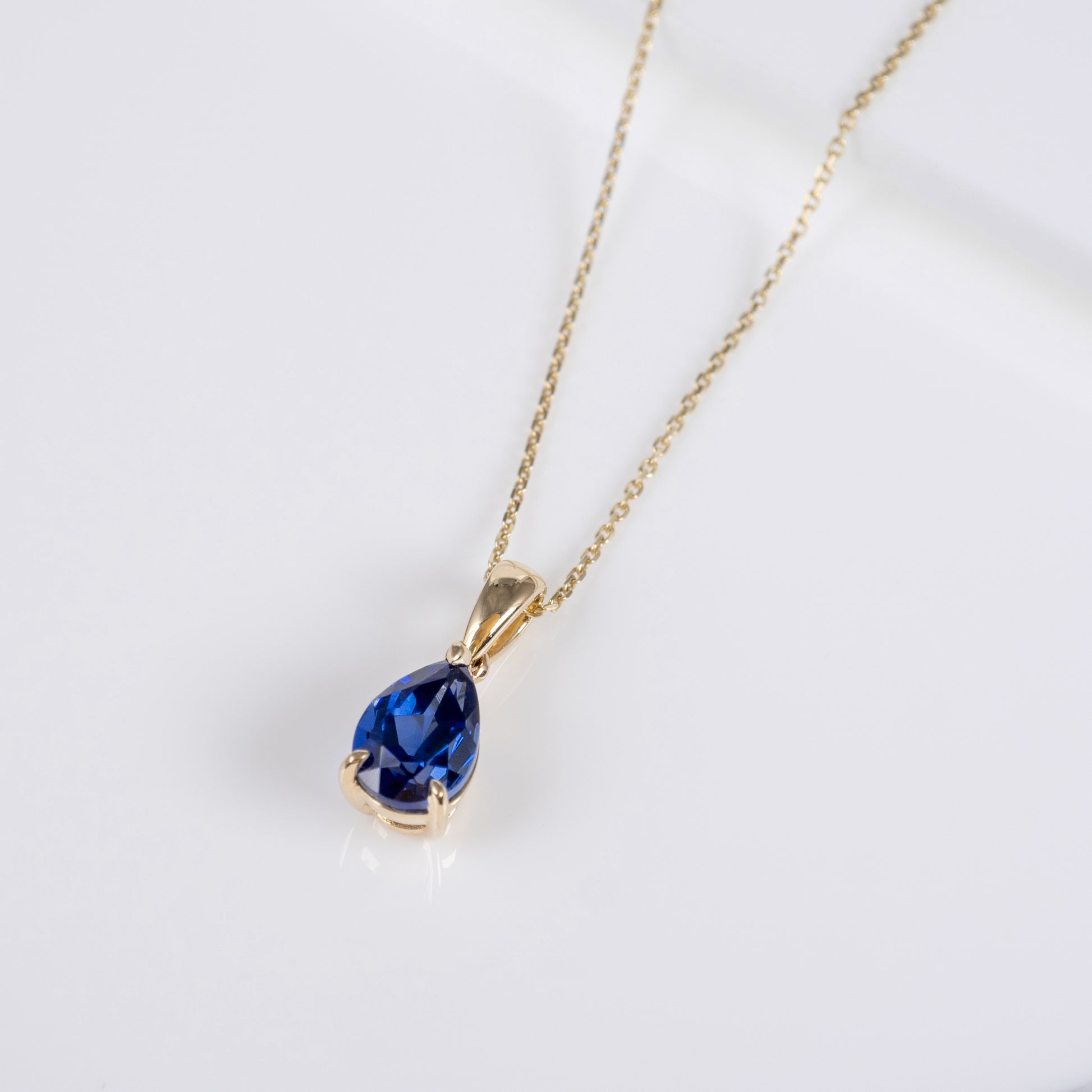 A gold chain necklace with a teardrop-shaped deep blue gemstone pendant, displayed on a clean white background.