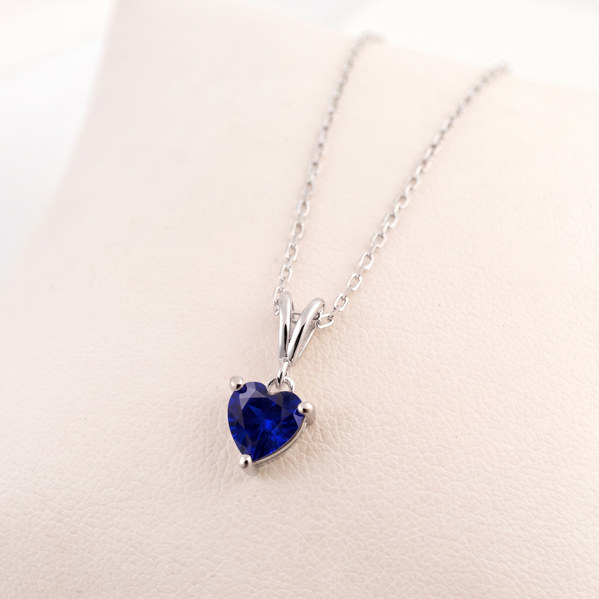 A silver heart-shaped pendant necklace with a deep royal blue gemstone, displayed on a light beige textured surface.