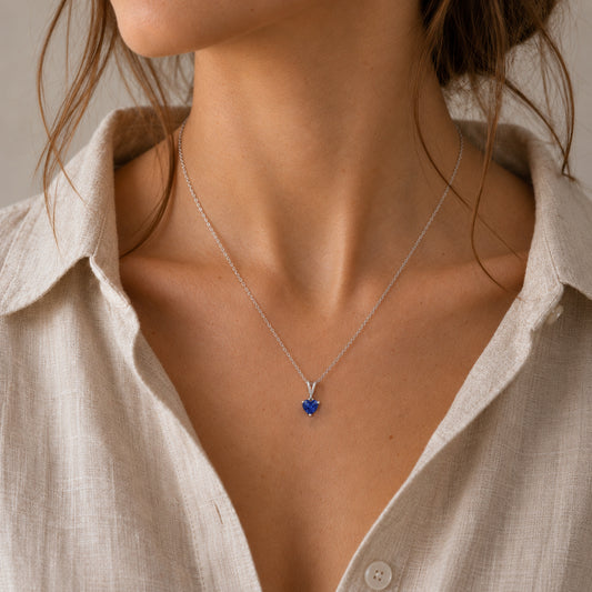 A close-up of a woman wearing a delicate silver chain necklace with a small blue heart-shaped pendant, styled with a light beige linen button-up shirt.
