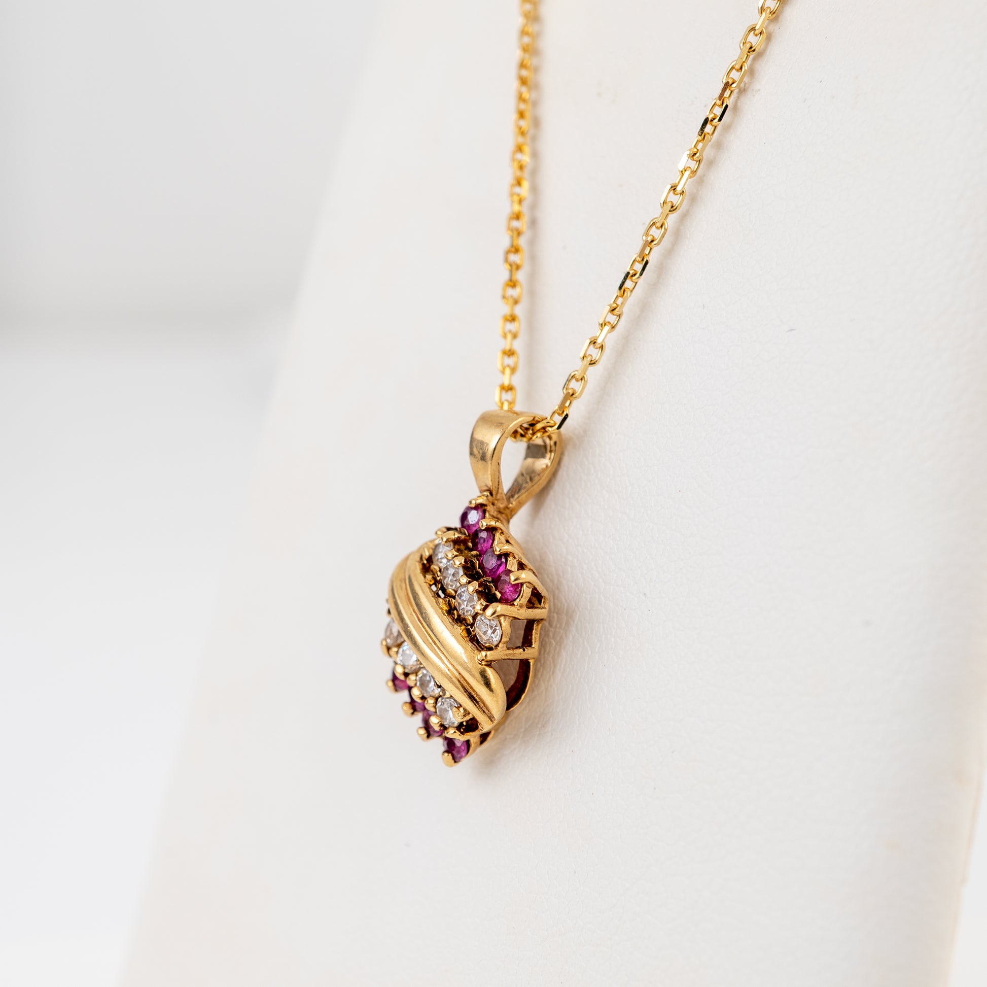 A gold pendant necklace with a wavy, openwork design set with small pinkish-red and clear stones, displayed on a white jewelry bust.