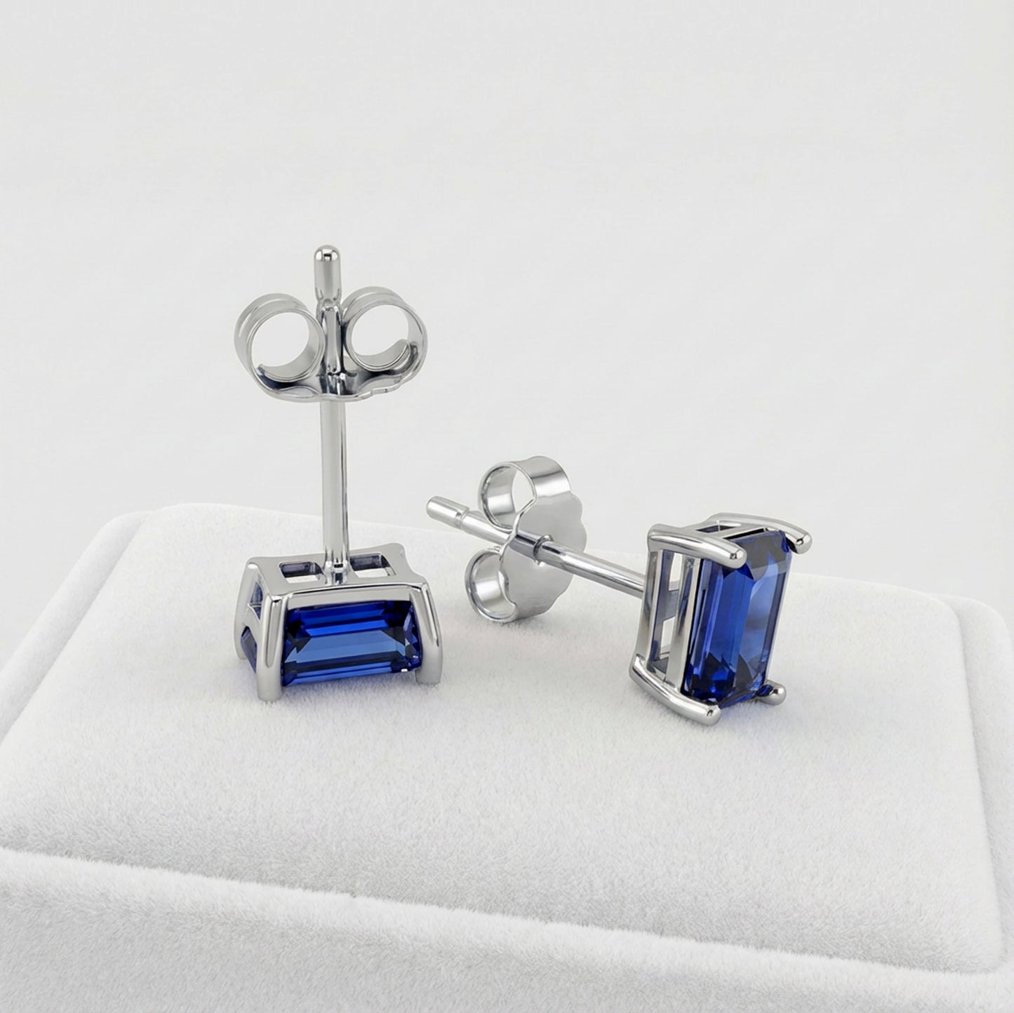 A pair of silver stud earrings with rectangular blue sapphire gemstones, displayed on a white jewelry cushion against a light gray background.