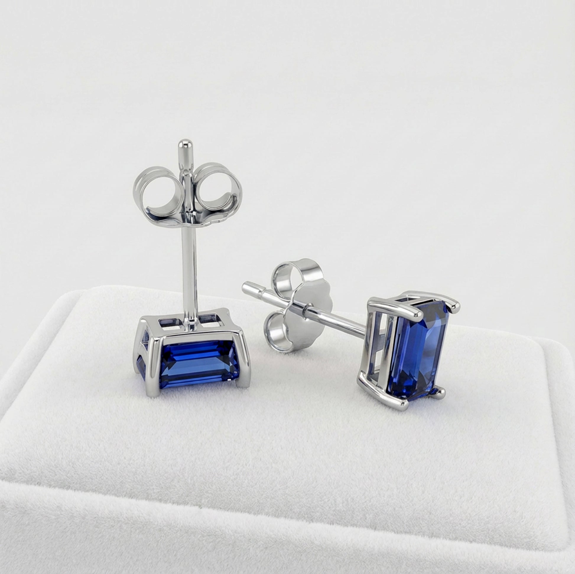 A pair of silver stud earrings with rectangular blue sapphire gemstones, displayed on a white jewelry cushion against a light gray background.