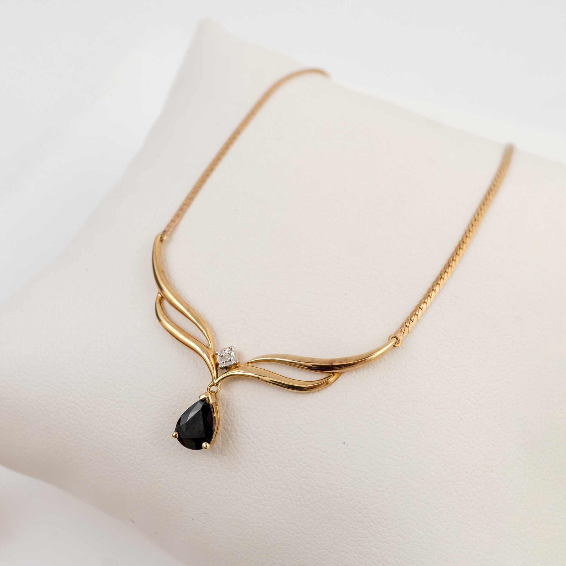 A gold necklace with a teardrop-shaped black gemstone pendant and a small clear accent stone, displayed on a cream-colored jewelry stand.