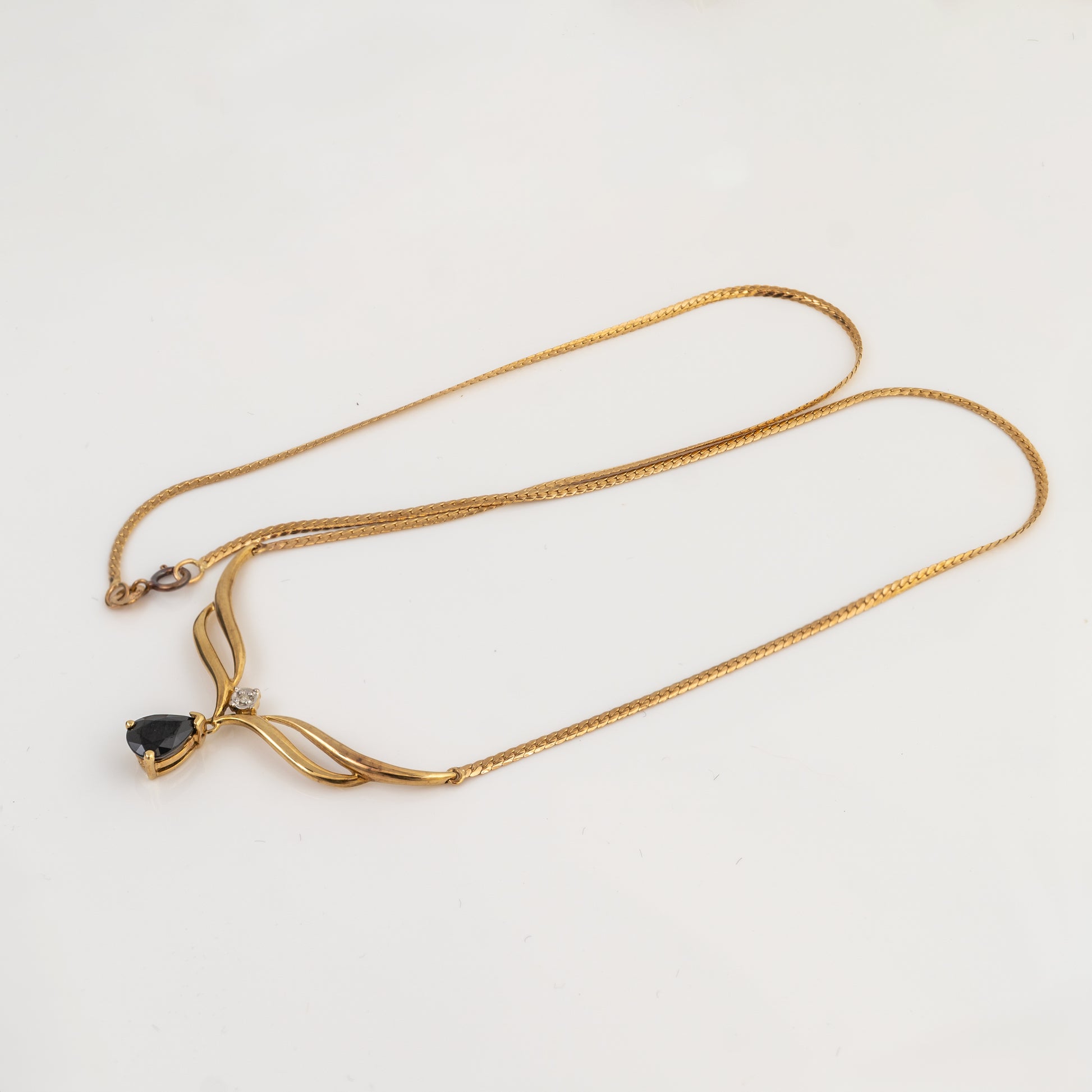 A gold chain necklace with a distinctive wavy pendant featuring a dark teardrop-shaped gemstone and a small clear accent stone, displayed on a clean white background.