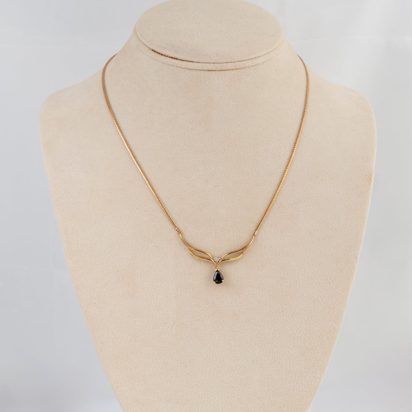 A delicate gold chain necklace with a small teardrop-shaped dark gemstone pendant, displayed on a beige fabric jewelry bust against a light gray background.